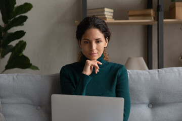 Wall Mural - Thinking by laptop. Thoughtful hispanic lady use laptop on sofa at home ponder on working problem read information on screen. Young female remote worker hold pc consider on electronic document