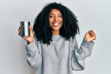 African american woman with afro hair holding canned food screaming proud, celebrating victory and success very excited with raised arm