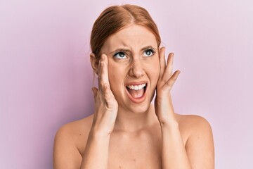 Wall Mural - Young irish woman standing topless stretching skin face with hands angry and mad screaming frustrated and furious, shouting with anger looking up.