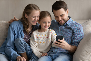 Wall Mural - Happy adorable small child girl using funny cellphone application with smiling young couple parents, enjoying playing mobile games, watching entertaining video in social network, shopping online.