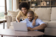 © fizkes - Happy young African couple focused on laptop screen watching movie, digital TV, sport match, relaxing at home. Husband and wife using computer together, pointing at screen, smiling