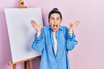 Poster - Young hispanic woman standing by painter easel stand celebrating crazy and amazed for success with arms raised and open eyes screaming excited. winner concept