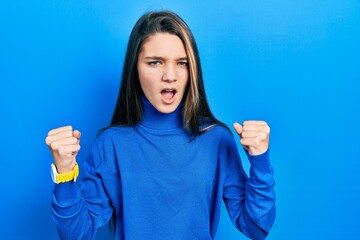 Poster - Young brunette girl wearing turtleneck sweater angry and mad raising fists frustrated and furious while shouting with anger. rage and aggressive concept.