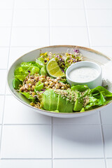 Green salad with avocado, sprouts, hemp seeds and yogurt dressing, white tile background.