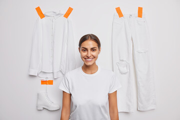 Wall Mural - Satisfied young woman in casual basic t shirt poses against white background with plastered shirt trousers and shoes sorts out wardrobe has good mood. Brunette female model poses near clothes