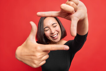 Wall Mural - Beautiful cheerful woman makes frame gesture smiles positively keeps eyes closed composes picture idea dressed in black jumper poses against vivid red background gazes at camera through hands