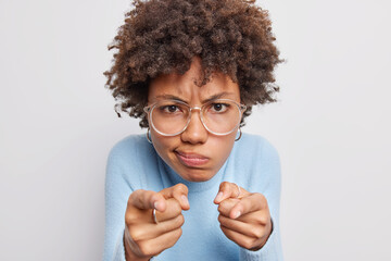 Wall Mural - Close up shot of displeased young curly woman looks angrily or with judgemental points index fingers at camera purses lips wears transparent glasses blue jumper isolated over white background.