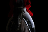 Fototapeta Konie - A rider in red jacket on horseback riding on dark background. Sportsman on black horse isolated on black background.