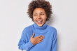 © wayhome.studio  - Portrait of cheerful curly haired woman smiles broadly keeps hand on chest cannot stop laughing at funny joke wears casual blue jumper isolated over white background. Positive human emotions concept