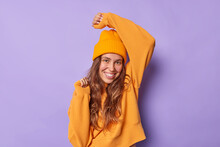 Photo Of Carefree Woman With Long Hair Smiles Happily Wears Casua Orange Jumper And Hat Keeps Arm Raised Dances With Rhythm Of Music Isolated Over Purple Background Has Fun. Happiness Concept