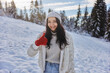 Happy young woman with a beautiful vivacious smile dressed warmly in winter clothes standing outdoors in front of forest giving thumbs up