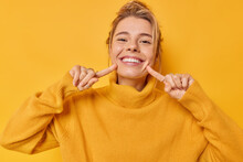 Portrait Of Cheerful Young Woman Smiles Broadly Indicates Index Fingers At Face Wears Casual Jumper Expresses Positive Emotions Isolated Over Vivid Yellow Background. Look At My Brilliant Smile
