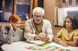 © pressmaster - Mature man shaking dice in hands over board game while playing it with his wife and their cute interracial granddaughter