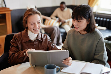 Wall Mural - Two young women sitting at the table and reading a book together, they preparing for homework