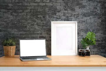 Laptop computer, empty picture frame, camera and houseplant on wooden table with brick wall.