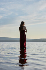 Wall Mural - Beautiful woman in light red dress stands in water of lake sea at sunset. Reflection of the girl and sky in water