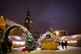 Fototapeta  - Winter evening on the main square of the city, street lit by evening lights, before Christmas, New Year. Christmas Fair. Catholic cathedral, church, tower. Snowy streets. Riga, Latvia.