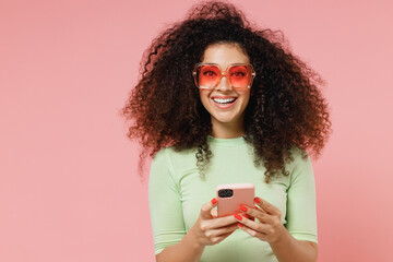 Wall Mural - Fascinating happy vivid exultant young curly latin woman 20s years old wears mint t-shirt sunglasses hold in hand use mobile cell phone isolated on plain pastel light pink background studio portrait.