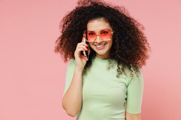 Wall Mural - Young curly latin woman 20s years old wears mint t-shirt sunglasses hold use talk on mobile cell phone conducting pleasant conversation isolated on plain pastel light pink background studio portrait.