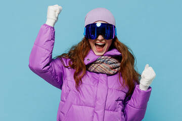 Wall Mural - Skier excited happy exultant caucasian woman in warm purple padded windbreaker jacket ski goggles mask spend extreme weekend in mountains do winner gesture isolated on plain blue background studio