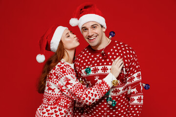 Wall Mural - Young lovely couple friends two man woman in sweater hat hold colorful garland kiss isolated on plain red background studio portrait. Happy New Year 2022 celebration merry ho x-mas holiday concept.