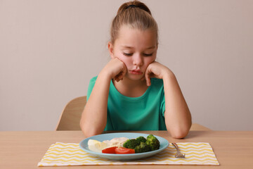 Cute little girl refusing to eat her breakfast at table on grey background