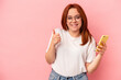 © Asier - Young caucasian woman holding a mobile phone isolated on pink background smiling and raising thumb up