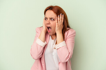 Young caucasian woman isolated on green background covering ears with hands trying not to hear too loud sound.