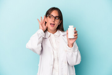 Wall Mural - Young pharmacist woman holding pills isolated on blue background trying to listening a gossip.