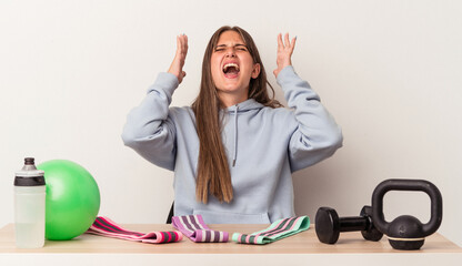 Wall Mural - Young caucasian woman sitting at a table with sport equipment isolated on white background screaming to the sky, looking up, frustrated.