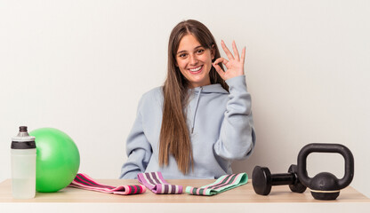 Wall Mural - Young caucasian woman sitting at a table with sport equipment isolated on white background cheerful and confident showing ok gesture.