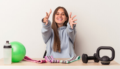 Wall Mural - Young caucasian woman sitting at a table with sport equipment isolated on white background feels confident giving a hug to the camera.