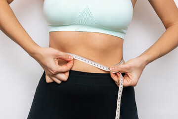 Wall Mural - Cropped shot of a young slender woman in sports clothes measuring her waist with a centimeter isolated on a white background. Close-up. Diet, sport and wellness concept