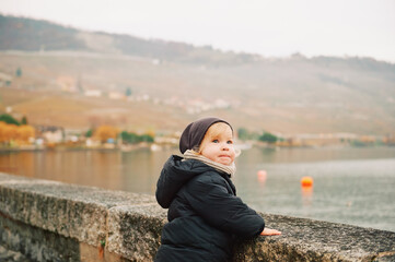 Wall Mural - Outdoor portrait of adorable toddler girl playing by the lake on a cold autumn day, wearing warm jacket and hat