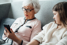 Grandmother Using Mobile Phone With Granddaughter On Sofa
