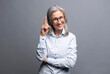 Have good idea. Smiling positive happy senior mature gray-haired businesswoman looks at the camera and holding index finger up, standing isolated on gray, came up with a solution, has solving of task
