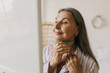 © shurkin_son - Beautiful mature grey-haired woman looking in mirror while doing facial massage with gua sha, taking care of her aging skin, neck area, enjoying moments on her own isolated on home interior background