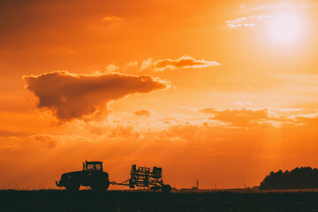 Wall Mural - Tractor Rides On Countryside Road. Beginning Of Agricultural Spring Season. Cultivator Pulled By A Tractor In Rural Field Landscape Under Sunny Summer Sunset Sunrise Sky. Backlit Dramatic Lighting