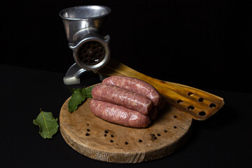 Canvas Print - Raw meat sausages on a wooden board on a black background. Homemade sausages in a natural casing next to a steel meat grinder. Meat products. Home cooking