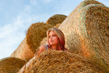 A Young Beautiful Blonde Girl With Blue Eyes And Snow-white Skin Sits On The Ground, Leaning On A Haystack, Looks Relaxed To The Side Enjoying The Rays Of The Setting Sun In Summer.