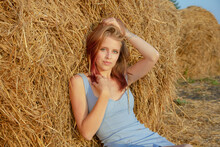 A Young Beautiful Blonde Girl With Blue Eyes And Snow-white Skin Sits On The Ground, Leaning On A Haystack, Looks Relaxed To The Side Enjoying The Rays Of The Setting Sun In Summer.