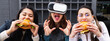 Horizontal banner or header portrait with young women sitting at restaurant eating burgers.  woman wears virtual reality glasses and bite a fictional hamburger. Real life versus virtual concept.
