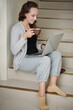 © DragonImages - Smiling young female entrepreneur sitting on steps at home, driking coffee and reading document on laptop screen