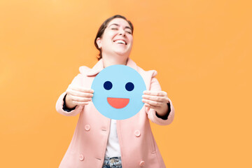 Happy girl holding piece of paper with blue emoji, isolated on orange background. Blue monday concept.