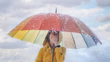 Happy Young Woman In Yellow Raincoat Under Rainbow Umbrella In Splashing Water