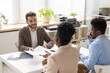© pressmaster - Smiling handsome young bearded lawyer in jacket sitting at table and giving documents to couple in office
