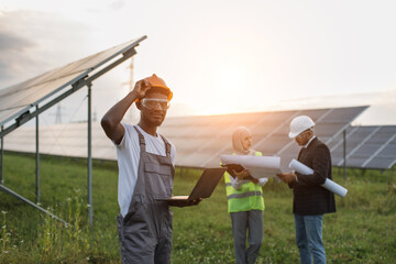 Wall Mural - African american technician in overalls looking at camera while standing at solar station with laptop in hands. Muslim woman and indian man examining blueprints on background.