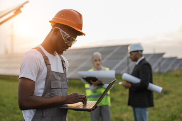 Wall Mural - African american man in yellow helmet and grey overalls typing on laptop while standing among rows of solar panels. Male and female inspectors standing behind and examining business plan.