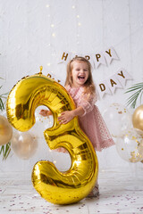 happy little blond girl in pink dress holding big gold foil balloon number and digit three 3