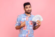 © khosrork - Young adult man with beard and pleasant appearance holding dollars bills and pointing at himself with pride, wearing blue casual style shirt . Indoor studio shot isolated on pink background.